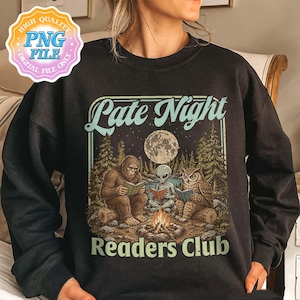 Op de afbeelding: Een zwart sweatshirt met lange mouwen en een grafische print van Bigfoot, een alien en een uil die rond een kampvuur zitten te lezen onder een volle maan in een bos. De tekst "Late Night Readers Club" staat in lichtblauw boven en onder.