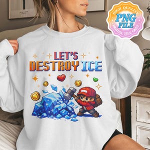 Könnte beinhalten: Weißes Sweatshirt mit dem Text "LET'S DESTROY ICE" in pixeligen orangefarbenen und blauen Buchstaben. Eine pixelige Grafik zeigt eine Figur, die mit einem Hammer einen blauen Eisblock zerschlägt, umgeben von Herzen und Münzen.
