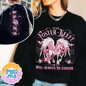 Può includere: Maglietta nera con una grafica di unicorno rosa e il testo "Team Foster-Keefe Will Always Be Cooler".