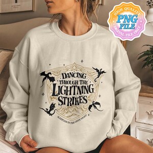 May include: Cream-colored sweatshirt with a graphic design featuring the text "Dancing Through The Lightning Strikes" in a stylized font. The design includes black silhouettes of dragons and a mountain landscape. The phrase "Sleepless in the Onyx Night" is also present.