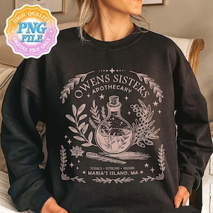 May include: A black long-sleeve sweatshirt featuring a light pink graphic print. The design displays a potion bottle, botanical elements, and the text "OWENS SISTERS APOTHECARY" along with "TONICS POTIONS ELIXIRS" and "MARIA'S ISLAND, MA" in a decorative frame.