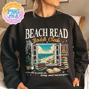 Op de afbeelding: Zwart sweatshirt met de tekst "BEACH READ Book Club" boven een illustratie van een open raam met uitzicht op een strand. Het ontwerp bevat stapels boeken en de zin "Just One More Chapter."