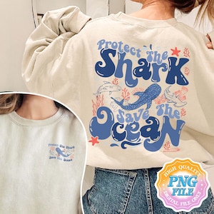 Puede incluir: Una sudadera beige con un estampado gráfico azul y rojo que dice "Protege al tiburón Salva el océano". El gráfico presenta un tiburón ballena, un tiburón martillo y arrecifes de coral.