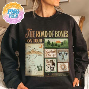 May include: A black crewneck sweatshirt featuring a graphic print titled The Road of Bones On Tour. The design includes a map with a dotted path, a forest scene, and rectangular stamps labeled Sunshine & Axe Eyes, Bloodaxe Crew, and The Owl's Inn with a skull and owl illustration. The text includes various locations and event details.