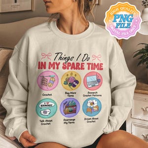 May include: Cream-colored sweatshirt featuring the text "Things I Do In My Spare Time" with illustrations of crochet-related activities. The design includes images of yarn, a laptop, and the words "Crochet," "Buy More Yarns," and "Research Crochet Patterns."