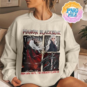 May include: A white sweatshirt with a graphic print of a woman with long white hair and a black cape. The text on the graphic reads "Manon Blackbeak, We are the Thirteen, From now until the darkness claims us".