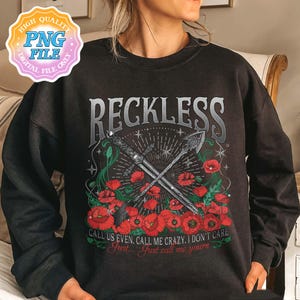 Puede incluir: Sudadera negra con un diseño gráfico que presenta un par de espadas cruzadas, flores rojas y la palabra "RECKLESS" en negrita. El texto debajo dice "CALL US EVEN. CALL ME CRAZY. I DON'T CARE Just... Just call me yours."