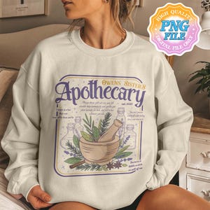 Peut inclure: Un sweat-shirt à col rond couleur crème avec une impression graphique portant le texte "Owens Sister's Apothecary est. 2025". Le motif représente un mortier et un pilon entourés d'herbes, de flacons en verre et de petites étoiles violettes et vertes.