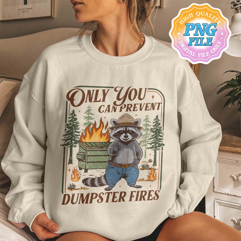 May include: Cream-colored sweatshirt with a graphic featuring a raccoon in a park ranger hat, standing in front of a dumpster fire. Text reads "Only You Can Prevent Dumpster Fires". The design includes trees and flames.