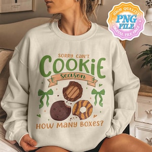 May include: Cream-colored sweatshirt with the text "Sorry, Can't. Cookie Season" in green and brown. The design includes illustrations of cookies and green bows. The phrase "How Many Boxes?" is also printed on the sweatshirt.