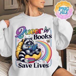 Puede incluir: Sudadera blanca con un gráfico colorido que presenta un mapache leyendo un libro, un arcoíris y el texto "Queer Books Save Lives". El mapache está sentado en un puf amarillo. Mariposas y libros también están en el diseño.