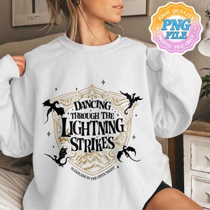 May include: White sweatshirt featuring a gold and black graphic with the text "Dancing Through The Lightning Strikes" and dragon silhouettes. The phrase "Sleepless in the Onyx Night" is also included. A "High Quality PNG File" digital file only sticker is in the upper right corner.
