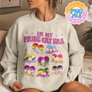 May include: Cream-colored sweatshirt with the text "IN MY PRIDE CAT ERA" in pink and purple. The design includes cat illustrations within rainbow and pride flag-themed hearts, representing various identities.