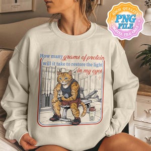 May include: Cream-colored sweatshirt with a humorous graphic of a cat lifting weights in a gym setting. The text reads, "How many grams of protein will it take to restore the light in my eyes." The design includes a red and blue border.