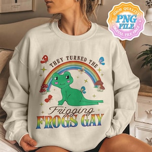 May include: Cream-colored sweatshirt with a whimsical design featuring a green frog under a rainbow. The text reads "THEY TURNED THE FRIGGING FROGS GAY" in colorful, playful lettering, with butterflies and mushrooms.