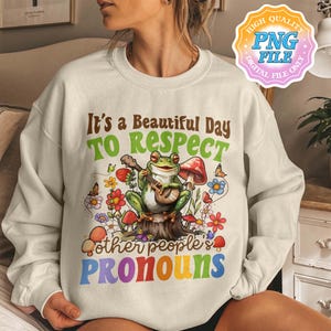 Può includere: Felpa color crema con la scritta "It's a Beautiful Day To Respect Other People's Pronouns". La grafica presenta una rana che suona il banjo su un ceppo d'albero, circondata da fiori e funghi. Un logo di file digitale si trova nell'angolo in alto a destra.