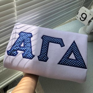 Puede incluir: Sudadera lila con las letras griegas ΑΓΔ en azul marino con lunares blancos. Las letras están bordadas en la tela. La sudadera está cerca de una ventana.