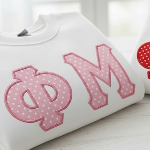May include: White sweatshirt with pink polka dot letters "ΦM". The letters are outlined in pink and have a light pink polka dot pattern. The sweatshirt is made of a soft material and is perfect for casual wear.