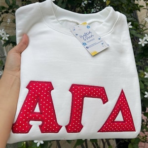 May include: A white sweatshirt with red, polka-dotted letters spelling out "AΓA". The letters are embroidered onto the front of the sweatshirt. A tag that says "Crafted & Decor" is attached to the sweatshirt.