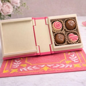 May include: A miniature, open, pink and cream-colored box of chocolates. The box contains four chocolates with different designs, including a swirl, sprinkles, and a heart shape. The box sits on a pink mat with a gold leaf design.