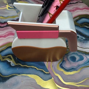 May include: A white and brown desk organizer shaped like a coffee pot, with a pink accent on the handle. The organizer holds several pens, including a black pen and a red pen with a pink pattern. The design is modern and functional.