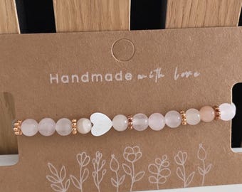 May include: A pink and rose gold beaded bracelet with a white heart charm. The bracelet is on a brown card with the text "Handmade with love".