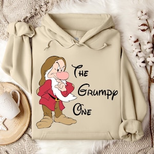May include: A beige hoodie featuring a cartoon image of Grumpy from Snow White. The character is depicted in a red coat and brown hair, with the text "The Grumpy One" in black script.