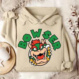 May include: A beige hoodie featuring a graphic of Bowser from the Super Mario video game series. The word "BOWSER" is printed in green block letters above the character's image. The character is green, yellow, and red.