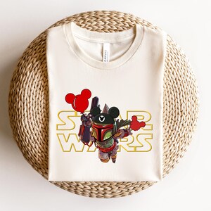 May include: Cream-colored t-shirt featuring a cartoon character in a Boba Fett helmet with a Mickey Mouse head. The words "STAR WARS" are in gold. The character is holding a weapon and a red balloon.