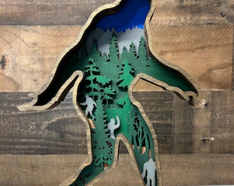 May include: A wooden cutout of Bigfoot walking through a forest. The cutout is layered to create a 3D effect. The forest is green with blue sky in the background.