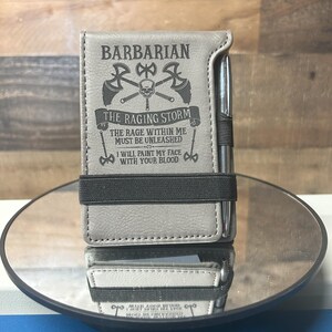 May include: A gray leather notebook with a pen holder and a black elastic band. The notebook has a design of crossed axes and a skull with the text "BARBARIAN THE RAGING STORM THE RAGE WITHIN ME MUST BE UNLEASHED I WILL PAINT MY FACE WITH YOUR BLOOD".
