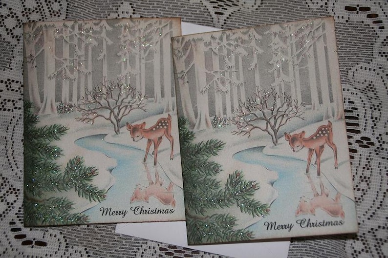 Peut inclure: Deux cartes de No&euml;l vintage repr&eacute;sentant une sc&egrave;ne de for&ecirc;t hivernale avec un faon pr&egrave;s d'un ruisseau. Les cartes pr&eacute;sentent un paysage enneig&eacute; avec des arbres et l'inscription "Merry Christmas". Les cartes sont d&eacute;cor&eacute;es de paillettes.