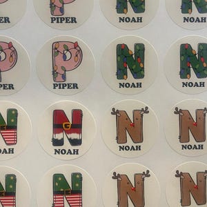 May include: A collection of round stickers featuring letters and names. The letters 'P' and 'N' are decorated with Christmas lights, Santa Claus belts, and reindeer antlers. The names 'Piper' and 'Noah' are printed below.