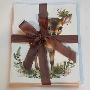 May include: A stack of blank greeting cards tied with a brown ribbon. The cards feature a watercolour illustration of a fawn wearing a wreath of holly and berries.