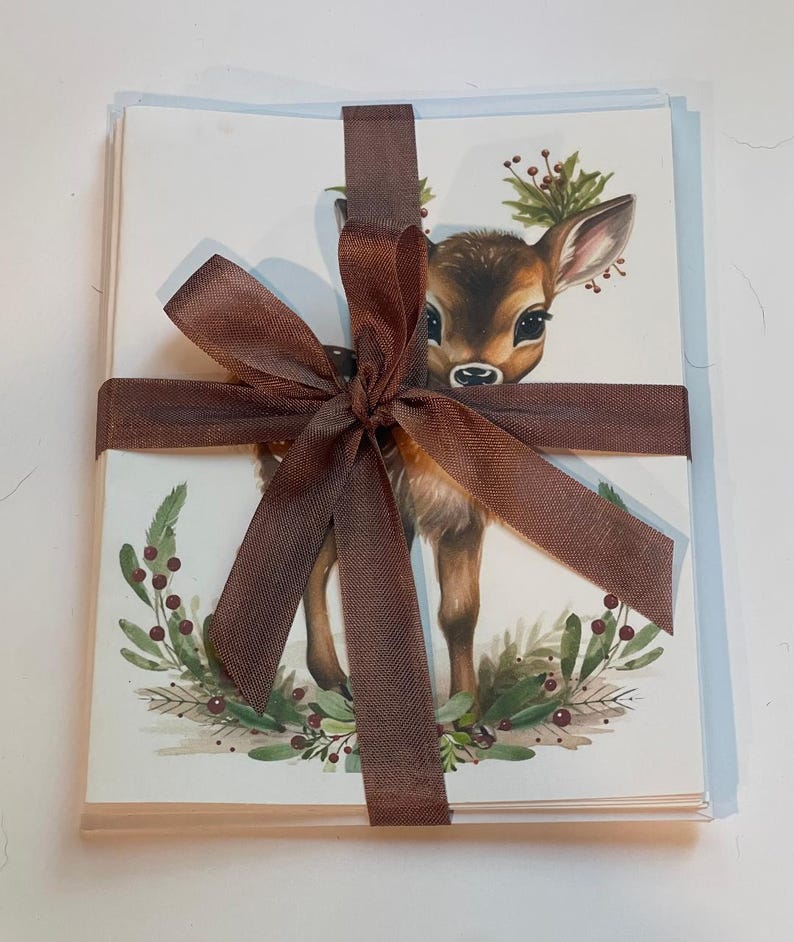 May include: A stack of blank greeting cards tied with a brown ribbon. The cards feature a watercolour illustration of a fawn wearing a wreath of holly and berries.