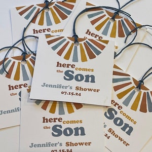 May include: White gift tags with a sunburst design in shades of brown, yellow, and blue. The text "here comes the Son" is printed on the tags, along with "Jennifer's Shower" and the date 07.15.24. Each tag has a dark blue string.