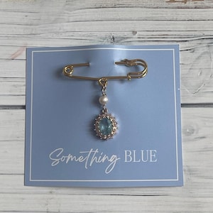 May include: A gold-colored safety pin with a dangling pendant. The pendant features a light blue gemstone surrounded by small clear stones and a white pearl. The pin is attached to a light blue card with the words "Something BLUE".