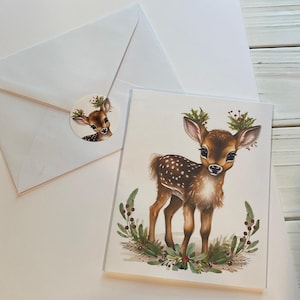 May include: A white envelope with a round sticker featuring a watercolour illustration of a fawn with a red berry wreath. The sticker is attached to the envelope. A white card with a watercolour illustration of a fawn with a red berry wreath is next to the envelope.