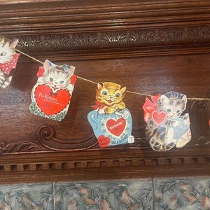 May include: A vintage Valentine's Day garland with five paper cutouts of cats. Each cutout features a different cat design with a red heart and a Valentine's Day message. The garland is strung with twine and is perfect for decorating for the holiday.