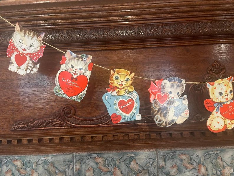 May include: A vintage Valentine's Day garland with five paper cutouts of cats. Each cutout features a different cat design with a red heart and a Valentine's Day message. The garland is strung with twine and is perfect for decorating for the holiday.