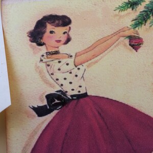 Op de afbeelding: Vintage kerstkaartillustratie van een vrouw die een kerstboom versiert. De vrouw draagt een witte top met polkadots, een donkerrode rok en een zwarte strik. De kerstboom heeft groene takken en rode ornamenten.
