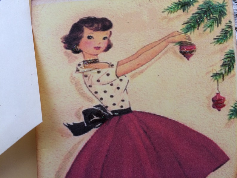Op de afbeelding: Vintage kerstkaartillustratie van een vrouw die een kerstboom versiert. De vrouw draagt een witte top met polkadots, een donkerrode rok en een zwarte strik. De kerstboom heeft groene takken en rode ornamenten.