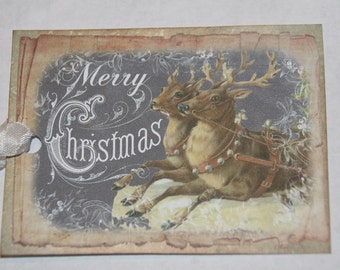 May include: A vintage-style Christmas gift tag with a gray background and the words "Merry Christmas" in white script. The tag features a drawing of two reindeer pulling a sleigh, with a white ribbon attached.