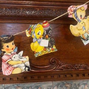May include: A vintage Easter garland with a string of paper cutouts. The garland features a girl holding a bunny, a chick and a black bird, and a chick with a basket of eggs. The garland is decorated with flowers and ribbons. The text on the garland reads "Easter Joy!"