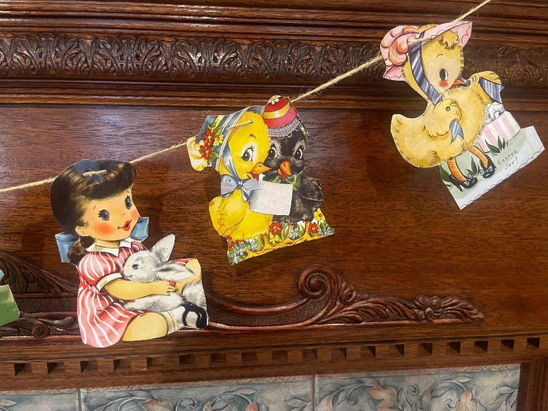 May include: A vintage Easter garland with a string of paper cutouts. The garland features a girl holding a bunny, a chick and a black bird, and a chick with a basket of eggs. The garland is decorated with flowers and ribbons. The text on the garland reads "Easter Joy!"
