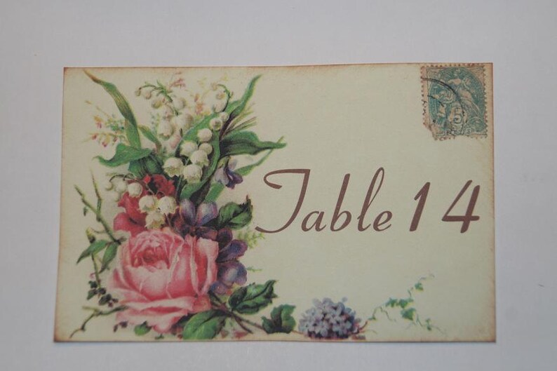May include: A vintage-style table number card with a floral design. The card features a bouquet of pink roses, white lilies of the valley, and purple flowers. The text "Table 14" is printed in brown cursive font.