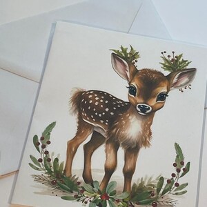 May include: A watercolour painting of a fawn with brown fur and white spots. The fawn is standing on a white background with a wreath of green leaves and red berries around it.