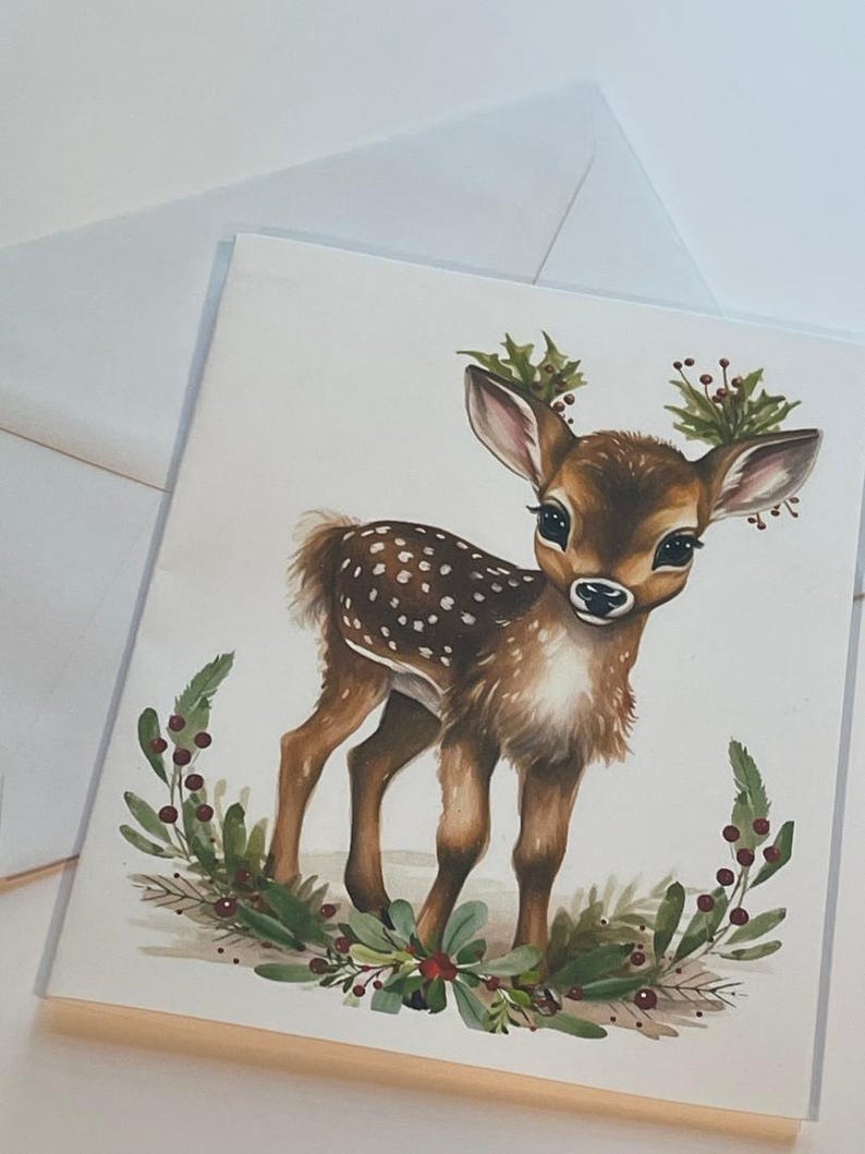 May include: A watercolour painting of a fawn with brown fur and white spots. The fawn is standing on a white background with a wreath of green leaves and red berries around it.