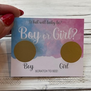 May include: A gender reveal scratch-off card with a watercolor background of blue and pink. The card reads "What will baby be? Boy or Girl?" with two gold scratch-off circles. Below the circles are the words "Boy" and "Girl".