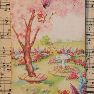 May include: A vintage-style illustration of a garden scene with a pink flowering tree, a birdhouse, a birdbath, and a watering can. The illustration is printed on a tag with a pink ribbon.
